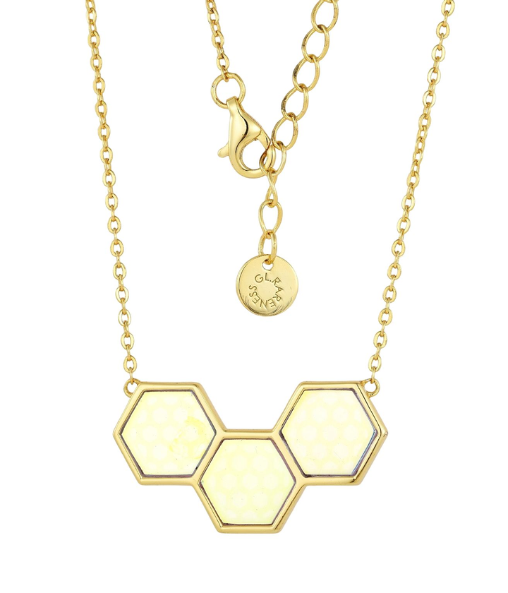 gold pendant necklace with three connected hexagons in a shallow chevron arrangement, each hexagon filled with pale honey-colored glass over a refracted honeycomb base pattern, fine cable chain with lobster clasp and brand disc

